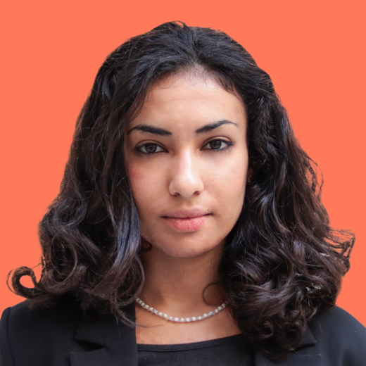 Photo de Kenza O.-Dj., une femme aux cheveux bruns et ondulés, portant une veste noire et un collier de perles, avec un arrière-plan orange uni. Elle a une expression neutre et confiante, avec un léger maquillage et un regard direct vers l'objectif.